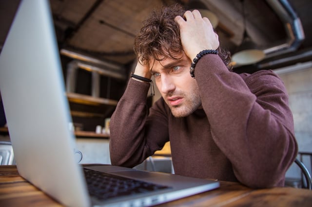 Stressful upset desperate handsome curly man in brown sweetshirt working using laptop and having headache.jpeg